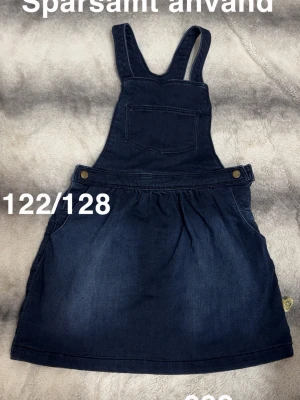 Mörkblå jeanskjol med hängslen - Snygg mörkblå jeanskjol med hängslen och bröstficka. Kjolen har två sidofickor och knappar vid midjan. Perfekt för en avslappnad och trendig look.🌸 Sparsamt använd. Strechigt och mjukt material. Storlek 122/128 