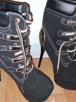 Svarta stövletter med gulddetaljer - Snygga svarta stövletter med höga stilettklackar och guldfärgade detaljer på hälen och snörningen. Skorna har vita kontrastsömmar och snörning framtill. Materialet är textil med inslag av syntet och metall. Perfekt för dig som vill sticka ut.