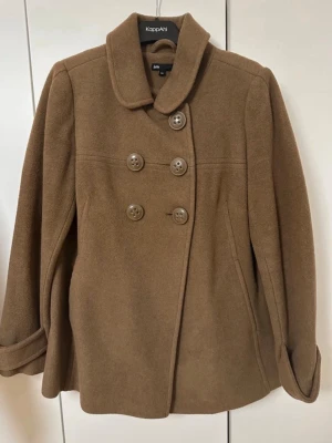 Brun ullkappa från jsfn - Stilren brun kappa från jsfn i ullblandning med dubbelknäppning framtill och dekorativa knappar bak. Kappan har bred krage, fickor i sidorna och snygga detaljer vid ärmsluten. Perfekt för kyliga dagar och ger en klassisk look.