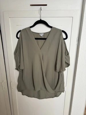 Beige omlottblus med öppna axlar - Snygg beige omlottblus med v-ringning och öppna axlar. Blusen har korta, fladdriga ärmar och är gjord i ett lätt, tunt tyg som ger ett luftigt intryck. Perfekt för en trendig och avslappnad stil.