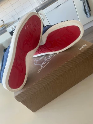 Blå Christian Louboutin sneakers - Snygga blå sneakers från Christian Louboutin med klassiska röda sulor och coola svarta nitar på tån. Skorna har en låg profil, vita kanter på sulan och är gjorda i tyg med läderdetaljer på insidan. Perfekta för dig som vill sticka ut med din stil.