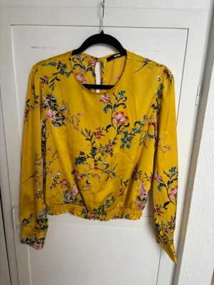 Gul blommig blus från Only - Snygg gul blus från Only med färgglatt blommönster i rosa, blått och grönt. Blusen har rund halsringning, lång ärm och resår i midjan för en avslappnad passform. Materialet känns lätt och luftigt, perfekt för en trendig look.