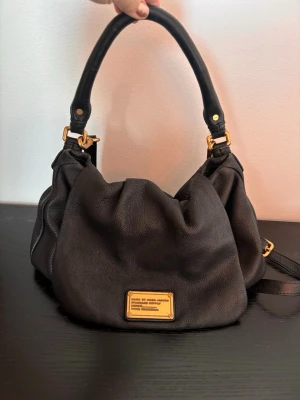 Svart läderväska Marc by Marc Jacobs - Snygg svart handväska i läder från Marc by Marc Jacobs. Väskan har en slouchy form, guldfärgade metalldetaljer och ett brett handtag. På framsidan sitter en metallbricka med logotyp. Perfekt för dig som gillar klassisk men cool stil.