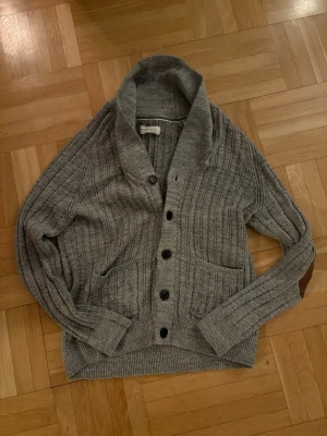 Grå ribbstickad kofta från Pier One - Snygg grå kofta från Pier One med ribbstickat mönster, sjalkrage och fem svarta knappar framtill. Två fickor framtill och breda muddar vid ärmslut och nederkant. Perfekt för lager på lager och chill stil.