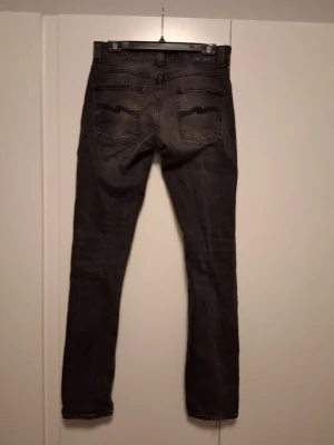 Nudie jeans Grim tim - Säljer ett par grå jeans från Nudie Jeans med klassisk femficksmodell och svarta vågdetaljer på bakfickorna. Jeansen har en rak passform och är tillverkade i denim med läderpatch i midjan. Perfekta för en avslappnad och stilren look. W29 L34