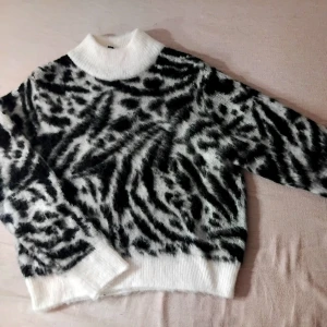 Stickad tröja - Säljer en fluffig stickad tröja med svartvitt mönster och hög krage. Tröjan har långa ärmar och ribbstickade muddar vid hals, ärmslut och nederkant. 