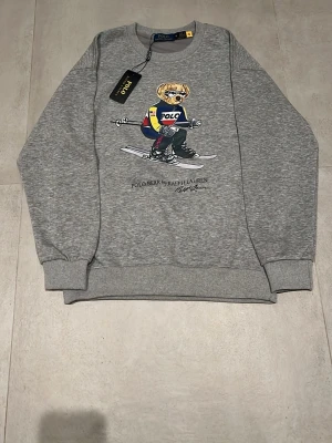 Grå Polo Bear hoodie från Ralph Lauren - Grå sweatshirt från Polo Ralph Lauren med ikoniska Polo Bear-trycket på bröstet. Rund halsringning, ribbade muddar och avslut. Tröjan har en cool björn med solglasögon och färgglad outfit på framsidan. Perfekt för en avslappnad och trendig look.