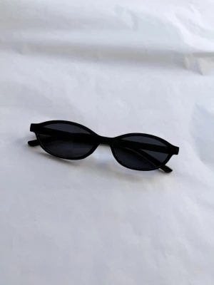 Svarta smala solglasögon - Snygga svarta solglasögon med smal, oval form och mörka glas. Ramen är i plast och ger en clean och modern vibe. Perfekta för dig som vill ha en stilren accessoar med edge.