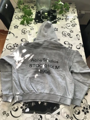 Grå hoodie Acne Studios Hoodie - Säljer en grå hoodie från Acne Studios med trycket 'Acne Studios STOCKHOLM 1996' på ryggen. Baggy passform med huva och ribbade muddar vid ärmar och midja. Tillverkad i mjuk bomullsblandning, perfekt för en avslappnad streetwear-look.