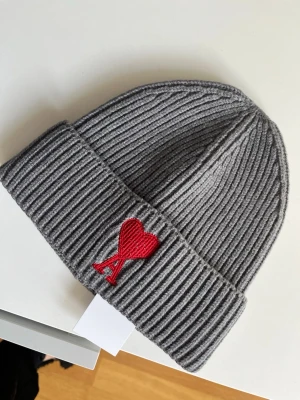 Grå ribbstickad mössa från Ami Paris - Säljer en grå ribbstickad mössa från Ami Paris med uppvikt kant och det ikoniska röda Ami de Coeur-hjärtat broderat framtill. Mössan är gjord i mjukt material och har en klassisk passform som passar perfekt till höst och vinter.