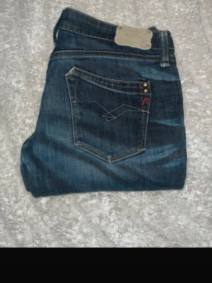 Mörkblå slim jeans från Replay - Snygga mörkblå slim jeans från Replay med klassisk femficksdesign och diskreta slitningar. Jeansen har kontrastsömmar, silverfärgade nitar och en liten röd detalj på bakfickan. Perfekta för dig som gillar en smal passform och tidlös stil.