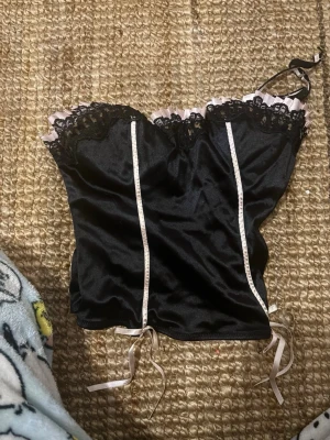 Svart body från Victoria's Secret - Snygg svart body från Victoria's Secret med spetsdetaljer upptill och ljusrosa volangkant. Smala axelband och dekorativa band framtill. Materialet är glansigt och känns lyxigt, perfekt för dig som vill ha något extra. 