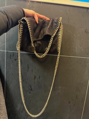 Svart axelväska med guld kedja - Cool svart axelväska i mjukt material med silverfärgade kedjor längs kanterna och som axelrem. Väskan har ett lock som viks över och dekorativa sömmar runtom. Perfekt för dig som gillar en edgy och trendig look.