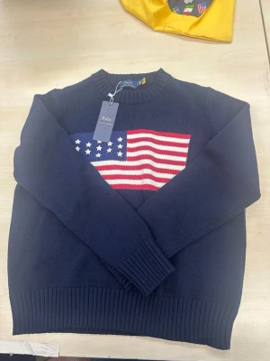 Mörkblå stickad tröja Polo Ralph Lauren - Mörkblå stickad tröja från Polo Ralph Lauren med amerikansk flagga i rött, vitt och blått på bröstet. Tröjan har rund halsringning och ribbstickade muddar vid ärmslut och nederkant. Perfekt för dig som gillar klassisk preppy stil. Tröjan är i helt nytt skick och kommer med taggen. Fråga bara om mer bilder om det behövs! Pris kan diskuteras vid snabba affärer.