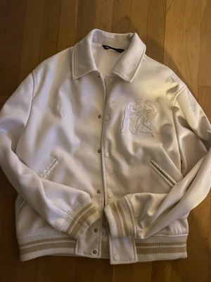 Vit bomberjacka från Zara - Snygg vit bomberjacka från Zara med broderad detalj på bröstet och ribbade muddar i vitt och beige. Jackan har tryckknappar framtill, två snedställda fickor och klassisk krage. look.