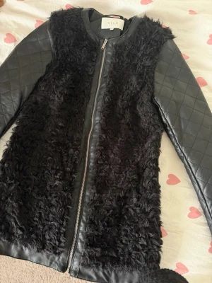 Svart pälsjacka från Vila XS - Snygg svart pälsjacka från Vila med quiltade ärmar i skinnimitation och fluffig päls framtill och baktill. Jackan har dragkedja framtill och en rak, modern passform. Perfekt för att hålla värmen under kalla dagar.