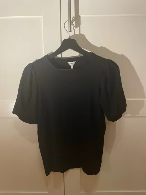 Svart topp med puffärm från Object - En svart topp från Object med puffiga korta ärmar och rund hals. Materialet är ribbat vilket ger en snygg struktur och toppen har en klassisk, stilren look som passar till det mesta. Perfekt för dig som gillar enkla men trendiga plagg.