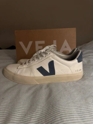 Vita Veja sneakers med blå detaljer - Snygga vita sneakers från Veja med blå V-logga på sidan och blå hälkappa med VEJA-tryck. Klassisk låg modell med snörning och platt sula. Tillverkade i skinn för en clean och stilren look.