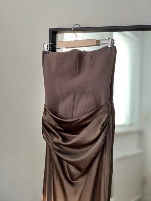House of CB: Brun axelbandslös långklänning - Brun axelbandslös långklänning från House or CB (Persephone Smoke Strapless Corset Maxi Dress) med draperad detalj framtill. Klänningen har en elegant, figurnära passform och slät yta som ger en lyxig känsla. Perfekt för dig som vill sticka ut med en stilren look. Denna klänning har använts en gång och har en mindre skråma på baksidan (se bild), i övrigt gott skick. Passar som balklänning, fest klänning eller bröllopsklänning.