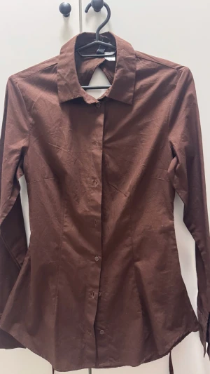Brun skjorta med öppen rygg - Snygg brun skjorta i med lång ärm, klassisk krage och knappar framtill, den har en öppen rygg och knytband. 
