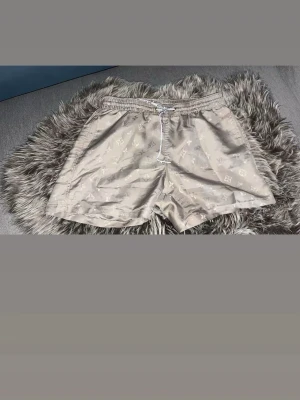 Beige shorts från Louis Vuitton  - Säljer ett par beige shorts från Louis Vuitton med klassiskt monogrammönster och vit snörning i midjan. Shortsen har elastisk midja och avslappnad passform, perfekta för sommardagar. Stilsäkert val för dig som gillar exklusiva detaljer.