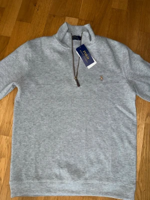 Grå halvzip från Polo Ralph Lauren - Snygg ljusgrå tröja från Polo Ralph Lauren med halv dragkedja och klassisk broderad logga på bröstet. Tillverkad i mjukt material och har ribbade muddar vid ärmslut och nederkant. Perfekt för en clean och stilren look.