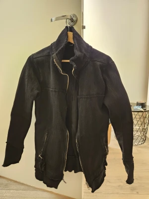 Svart cardigan jacka - Säljer en svart jacka med råa kanter och asymmetrisk dragkedja. Jackan har hög krage, två fickor med dragkedja och en lite längre, avslappnad passform. Använd endast 3-4 gånger och vid det tillfället gick dragkedjan sönder, som nu inte fungerar.