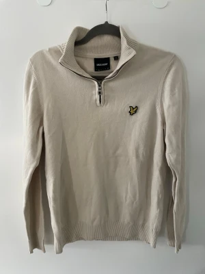 Beige halvzip tröja från Lyle & Scott - Snygg beige tröja från Lyle & Scott med halvzip och hög krage. Tröjan har ribbade muddar och det klassiska gula logomärket på bröstet. Perfekt för dig som gillar stilrena och tidlösa plagg. Materialet är mjukt och bekvämt.