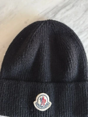 Svart ribbstickad mössa från Moncler - Snygg svart ribbstickad mössa från Moncler med uppvikt kant och klassisk Moncler-logga framtill. Tillverkad i mjukt material som håller värmen och ger en clean look. Perfekt accessoar för kyliga dagar.