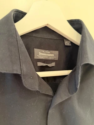Svart skjorta från Dressmann - Klassisk svart skjorta från Dressmann i bomull med stretch. Skjortan har lång ärm, knappar framtill och en stilren krage. Perfekt för dig som gillar en enkel och clean look.