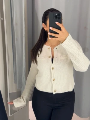 Vit ribbstickad kofta med guldknappar - Snygg vit ribbstickad kofta med guldfärgade knappar och rund halsringning. Kortare modell med dekorficka på bröstet och långa ärmar. Perfekt att styla med jeans eller kjol för en trendig look.