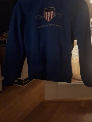 Blå hoodie från Gant med tryck - Säljer en blå hoodie från Gant med tryck på bröstet och ett stort Gant-emblem. Hoodien har huva och magficka, samt ribbade muddar vid ärmslut och nederkant. Perfekt för dig som gillar sportig och avslappnad stil.