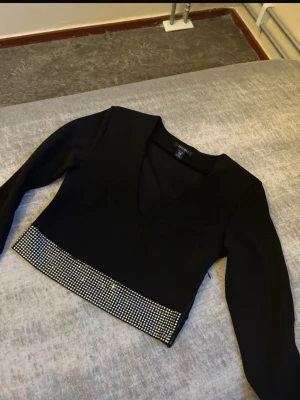 Svart croppad topp med paljetter - Säljer en svart croppad topp från Amisu med v-ringning och långa ärmar. Toppen har en snygg paljettkant längst ner som ger extra bling. Perfekt för dig som gillar att sticka ut och vill ha något trendigt i garderoben.