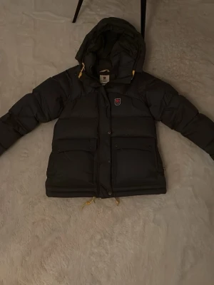 Mörkgrå pufferjacka från Fjällräven - Säljer en mörkgrå pufferjacka från Fjällräven med gula detaljer och huva. Jackan har två stora fickor framtill, dragsko i nederkant och klassisk Fjällräven-logga på bröstet. Perfekt för kalla vinterdagar och riktigt skön att bära.💞💞