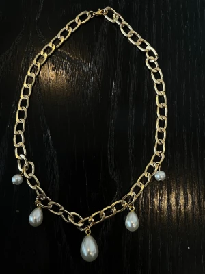 Guldfärgat halsband med pärldroppar - Snyggt halsband med grov guldfärgad kedja och fem hängande vita pärldroppar. Kedjan har en modern, chunky look och pärlorna ger en elegant touch. Perfekt för att lyfta vilken outfit som helst.