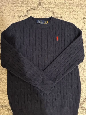 Mörkblå stickad tröja Polo Ralph Lauren - Kabelstickad mörkblå tröja från Polo Ralph Lauren med rund halsringning och den klassiska röda logotypen broderad på bröstet. Tröjan har ribbade muddar vid ärmslut och nederkant. Perfekt för lager på lager och snygg till jeans. Använde den 2 gånger och sen har den bara varit i garderoben. Den sitter lite dock lite tight och därför rekommenderar jag den för folk med storlek S.