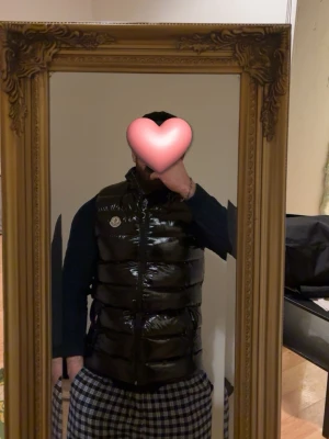 Svart dunväst från Moncler - Snygg svart dunväst från Moncler med glansig finish och klassisk logga på bröstet. Västen har hög krage, två framfickor med dragkedja och tydlig Moncler-text på insidan av dragkedjan. Perfekt för lager-på-lager och streetwear-stil.