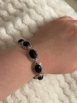 Silverfärgat armband  - Snyggt armband i silverfärg med dekorativa svarta pärlor och detaljerade metallinslag. Armbandet har en klassisk design med rundade former och passar perfekt för dig som gillar stilrena accessoarer med en twist.