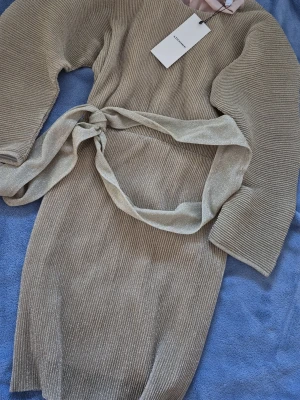 Glittrig beige klänning från Adoore - Supersnygg glittrig beige klänning från Adoore med ribbad struktur och matchande bälte i midjan. Klänningen har lång ärm och en elegant, figurnära passform som ger en lyxig känsla. Perfekt för dig som vill sticka ut med stil.