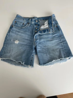 Levi's blå jeansshorts med rå kant - Säljer ett par klassiska blå jeansshorts från Levi's Premium med hög midja, knappar i gylfen och råa, fransiga benslut. Shortsen har en liten slitning på ena benet och femficksmodell. Perfekta till sommaren!
