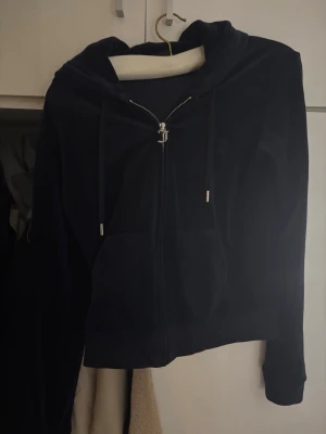 Mörkblå velour hoodie från Juicy Couture - Snygg mörkblå hoodie i mjuk velour från Juicy Couture. Dragkedja framtill, två fickor och justerbar huva med snören. Klassisk JC-logga broderad på bröstet. Perfekt för en chill och trendig look.