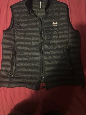 Svart dunväst från Colmar - Snygg svart dunväst från Colmar med quiltad design och dragkedja framtill. Västen har två praktiska fickor med dragkedja och en liten logga på bröstet. Perfekt att slänga över en hoodie eller tröja för extra värme och stil.