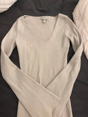 Beige ribbstickad klänning H&M - Säljer en beige ribbstickad klänning från H&M med lång ärm och djup rundad urringning. Klänningen är figurnära och har en mjuk, stretchig känsla . Enkel och stilren design som är lätt att matcha.