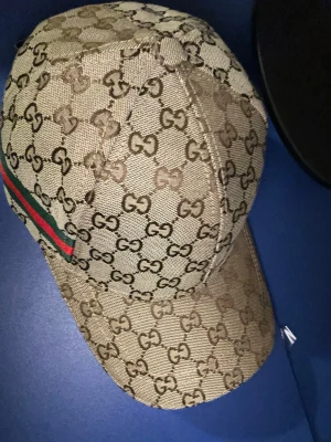 Beige Gucci keps med GG-mönster - Säljer en beige keps från Gucci med det klassiska GG-mönstret över hela kepsen. På sidan finns en grön och röd rand som signaturdetalj. Kepsen har böjd skärm och är tillverkad i slitstarkt bomullsmaterial. Perfekt för dig som vill ha en ikonisk och trendig accessoar.