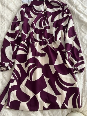 Mönstrad lila och vit klänning - Kort klänning med abstrakt mönster i lila och vitt. Klänningen har långa ärmar, markerad midja med knytband och rund halsringning. Perfekt för dig som gillar färg och unika prints. XXS, ny med etiketter 