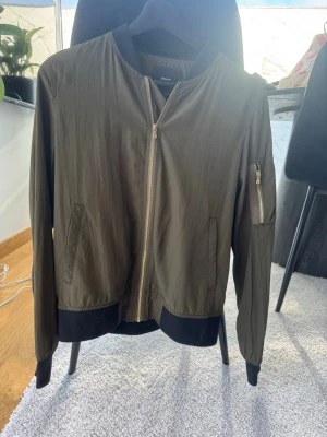 Olivgrön bomberjacka med gulddragkedja - Snygg olivgrön bomberjacka frånZara med guldfärgad dragkedja framtill och på ärmen. Jackan har svarta muddar vid ärmslut, krage och nederkant samt två fickor fram. Tillverkad i lätt polyester för en clean look och skön passform.