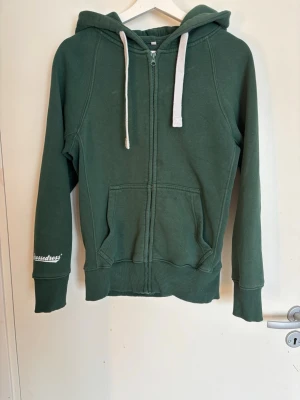 Russemurch hodie grön - Mörkgrön hoodie med dragkedja från russemurch . Har vita dragsnören i huvan, två stora fickor framtill och ribbade muddar vid ärmar och nederkant. Broderad logga på ena ärmen. Perfekt för en chill och avslappnad stil.