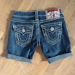 W23 True Religion shorts vintage y2k m vita sömmar - Vintage avklippta jeans till shorts från True Religion. Tydliga vita kontrastsömmar och stora bakfickor med dekorativa knappar. Svåra att få tag på! Klassisk femficksmodell och rakt snitt. Märkespatch bak i rött och vitt. Perfekta för dig som gillar Y2K. Storlek w23 (23 tum i midjan) motsvarar ungefär strl 32/xs. 