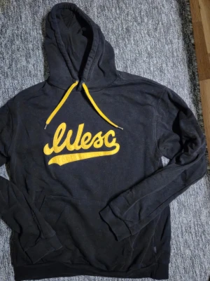 Svart hoodie med gul wesc-logga - Cool svart hoodie från Wesc med stor gul broderad logga på bröstet och matchande gula snören i huvan. Klassisk passform med känguruficka och ribbade muddar. Perfekt för dig som gillar streetwear och vill sticka ut med färg. STORLEK LARGE, FINNS I HÄSSELBY 