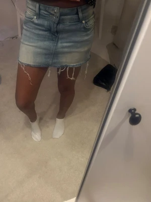 Ljusblå jeanskjol med fransar - Trendig ljusblå jeanskjol med råa fransar längst ner och klassiska fem fickor. Kjolen har en kort passform och stängs med dragkedja och knapp framtill. Perfekt för dig som gillar en avslappnad och cool stil.